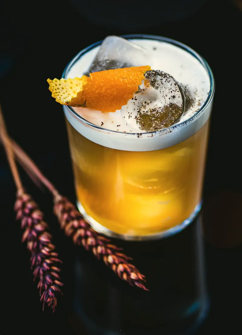 Drink amarelo com espuma e casca de laranja em copo baixo sobre fundo escuro