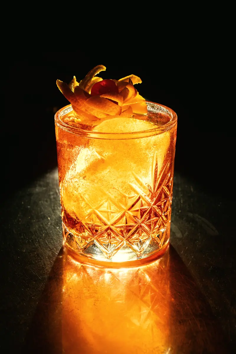 Drink âmbar com gelo e casca de laranja em copo de cristal lapidado, iluminado por luz quente lateral sobre fundo escuro